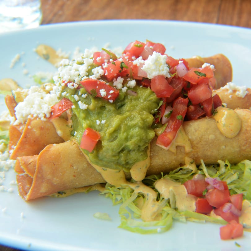 Crispy taquitos topped with guacamole, diced tomatoes, crumbled cheese, and a creamy sauce.