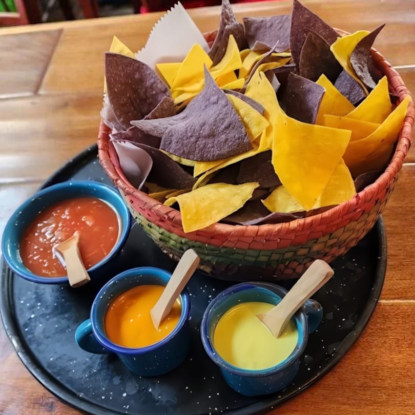 A colorful basket of yellow and blue corn tortilla chips with three dipping sauces on a black tray.